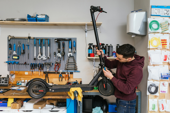 Mecánico de bicicletas ajustando un scooter eléctrico en la parte inferior del manillar cerca de la unión con la rueda, mientras está colocado en el banco de trabajo, con herramientas en el fondo de su taller.